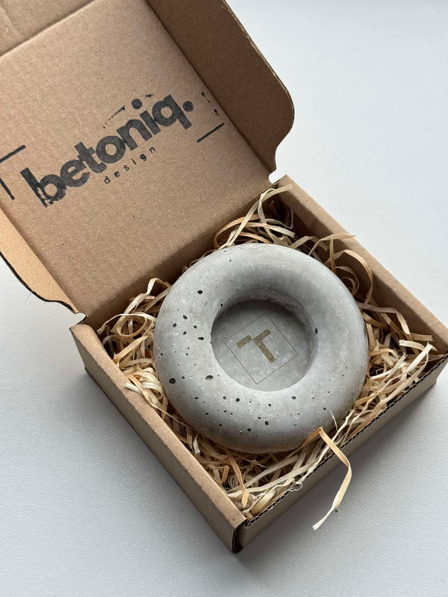 Děkuji za poslední fotku!

Na této fotce vidíme:

Otevřenou kartonovou krabičku s logem "betoniq design" na víku.

Uvnitř krabičky, na výplni z dřevěné vlny, je položený kulatý betonový svícen nebo dekorace ve tvaru prstence/věnce.

Uprostřed prstence je zobrazeno logo Betoniq (písmeno "T").

Zde je návrh na alt text:

Alt text: Kulatý betonový svícen nebo dekorace ve tvaru prstence s logem Betoniq uprostřed, balený v otevřené kartonové krabičce s logem Betoniq design a dřevěnou vlnou.