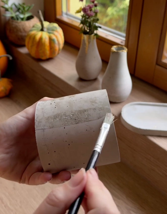 Ručně vyráběná betonová nádoba v procesu finální úpravy. Jemné detaily a precizní ruční práce dodávají produktu jedinečný minimalistický design. V pozadí podzimní dekorace a další betonové vázy se zlatými akcenty.