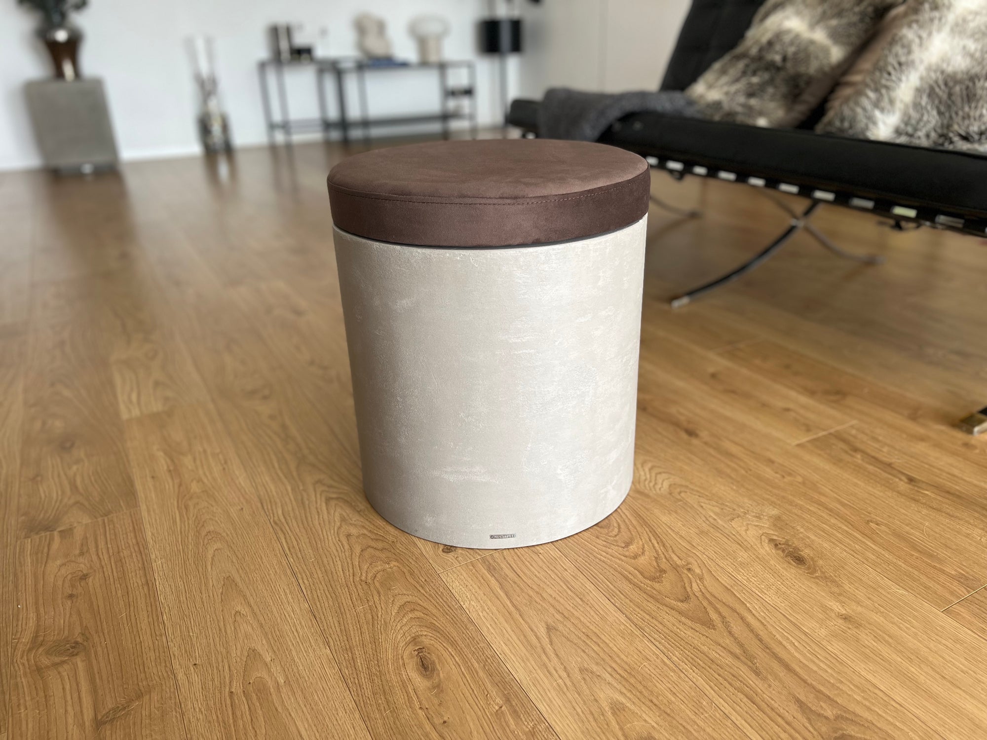 Mikrobetonový taburet se sedákem v hnědé barvě, umístěný na dřevěné podlaze v moderním obývacím pokoji.