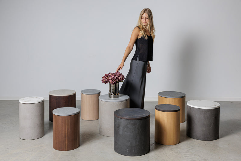 Noční stolky a odkládací stolky z mikrobetonu a dřeva, vyráběné v Evropě, v různých barvách a povrchových úpravách, zobrazené v moderním interiéru s ženou, která interaguje s nábytkem. Minimalistický a funkční design, ideální pro současnou dekoraci.