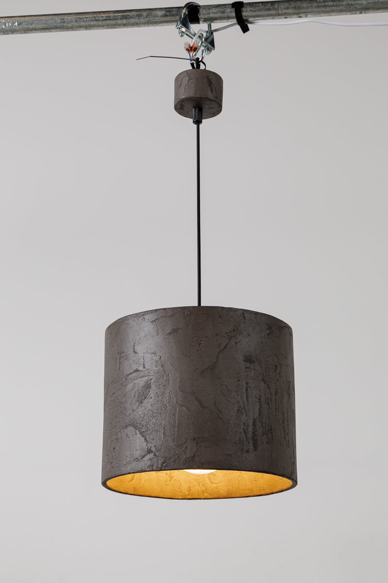 Moderní závěsná betonová lampa s cylindrickým stínidlem a teplým zlatým vnitřkem, zavěšená na černém kabelu, umístěná na minimalistickém bílém pozadí