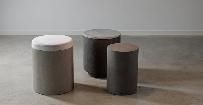 Skupina mikrobetonových odkládacích stolků a taburetu s polštářem v moderním interiéru. Představuje válcové tvary v šedých tónech a jeden závěsný prvek.