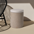 Světle béžový mikrobetonový odkládací stolek s kulatým bílým podsedákem, stojící na béžové podlaze se stínem vedle tmavé designové židle, ideální jako stylová taburetka nebo noční stolek.